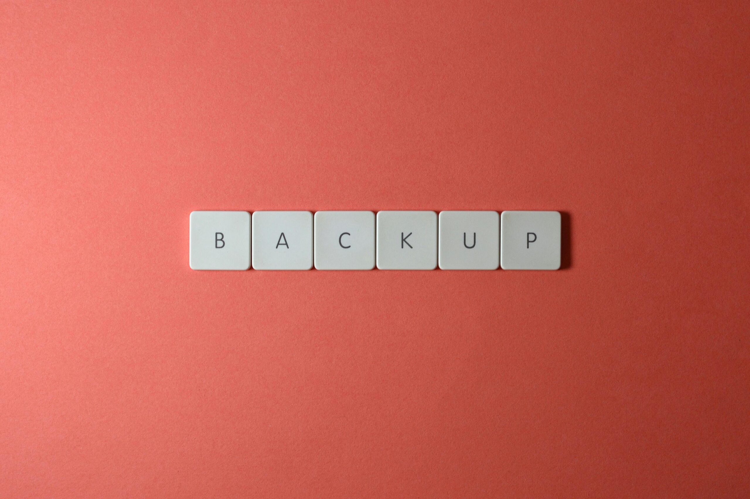 Free Close-Up Of Keyboard Keys Spelling 'Backup' Placed On A Coral-Colored Surface. Stock Photo